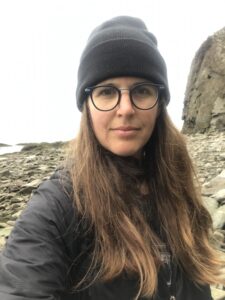 Image of Ashley Watson. She is on a rocky beach and is wearing a hat and glasses.